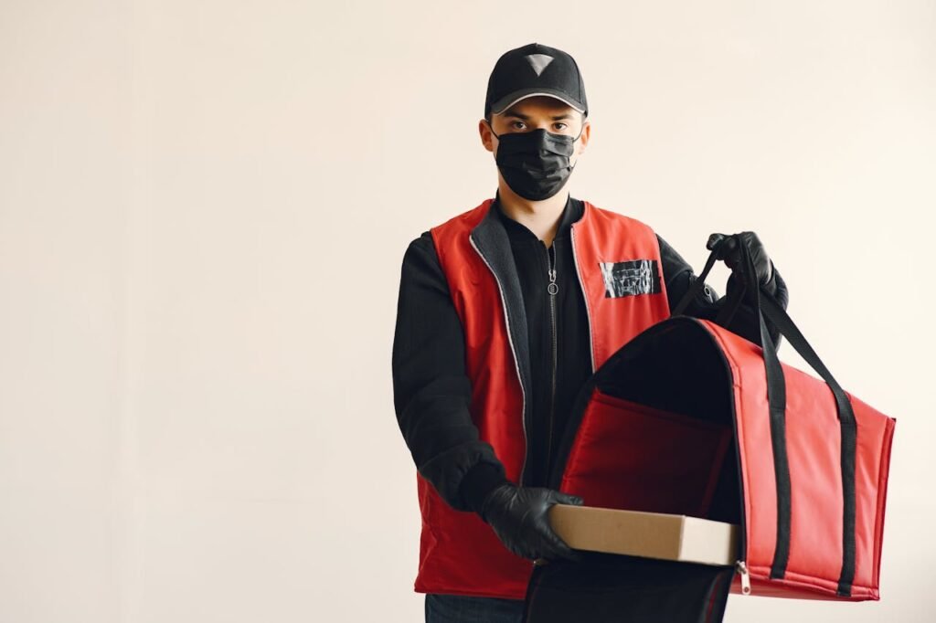 Delivery man wearing a mask and gloves with a red thermal bag and box indoors.